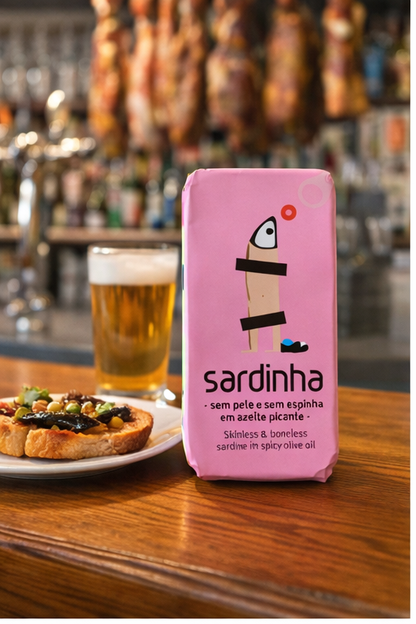 A can of Sardinha Skinless and Boneless Sardines in Spicy Olive Oil sits on a wooden bar next to a glass of beer and a small plate, with hanging cured meats and shelves blurred in the background.