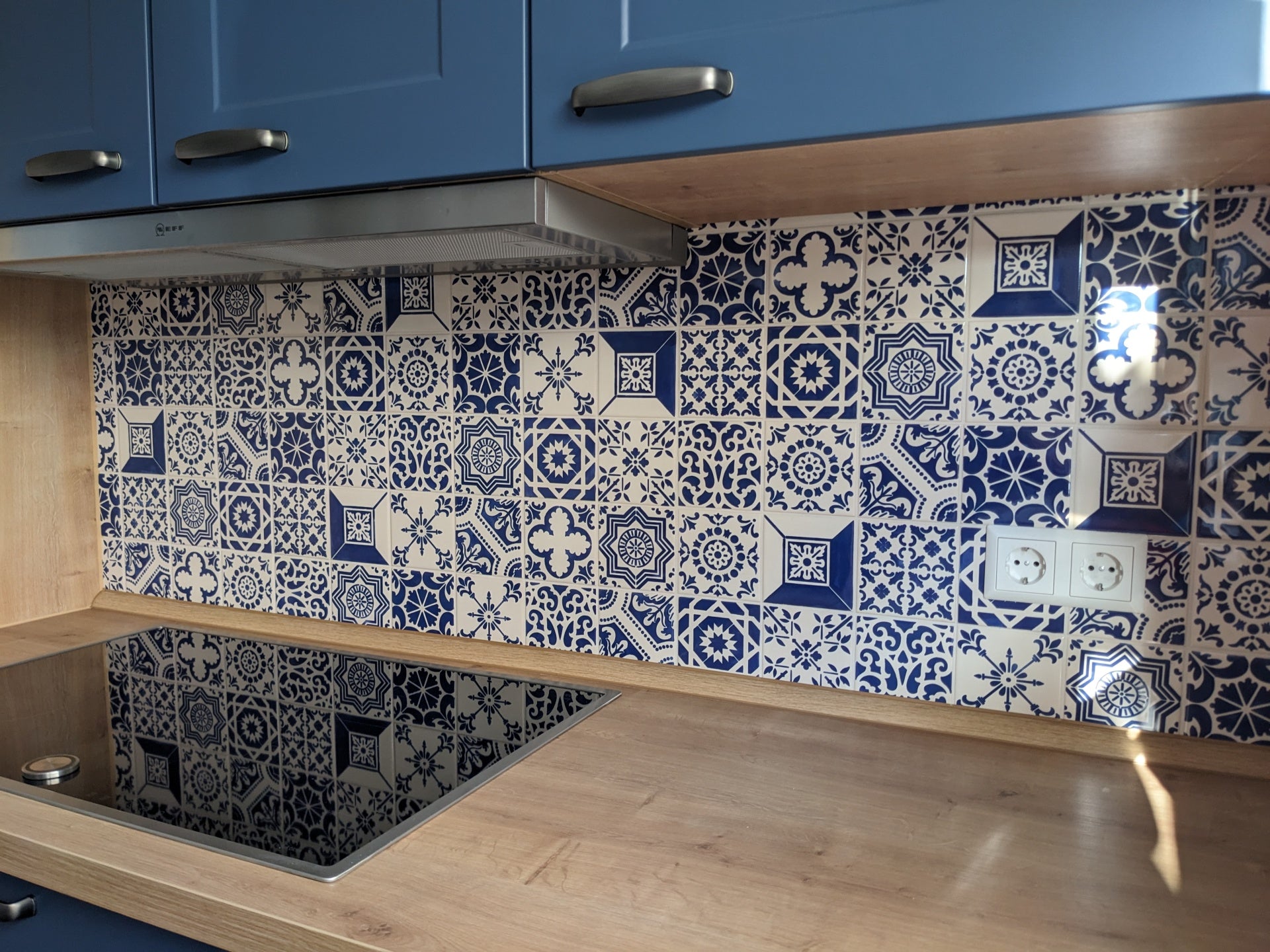A modern kitchen featuring blue cabinets, a wooden countertop, a black electric stove, and a TilePassion Hand Painted Splashback Set of Azulejo Tiles with blue and white patterns. Two electrical outlets are on the right side of the splashback.