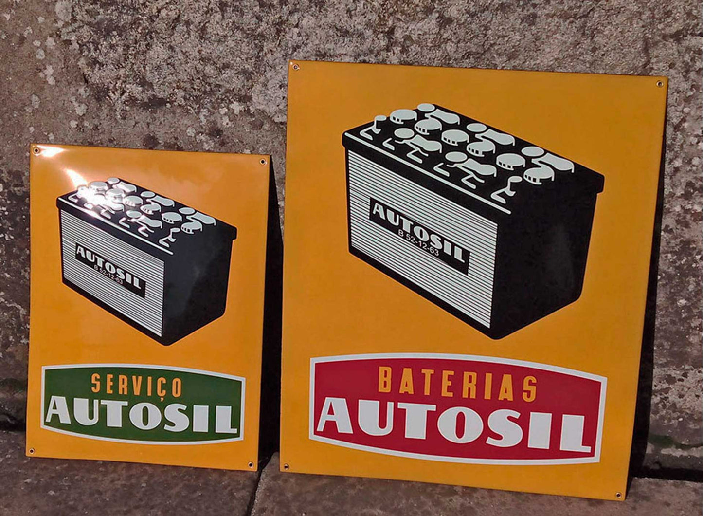 Two yellow Square Enamel Signs & Plaques by Luis Filipe feature car battery illustrations: one reads SERVIÇO AUTOSIL, the other BATERIAS AUTOSIL. Both vitrified enamel plaques are displayed against a stone background.