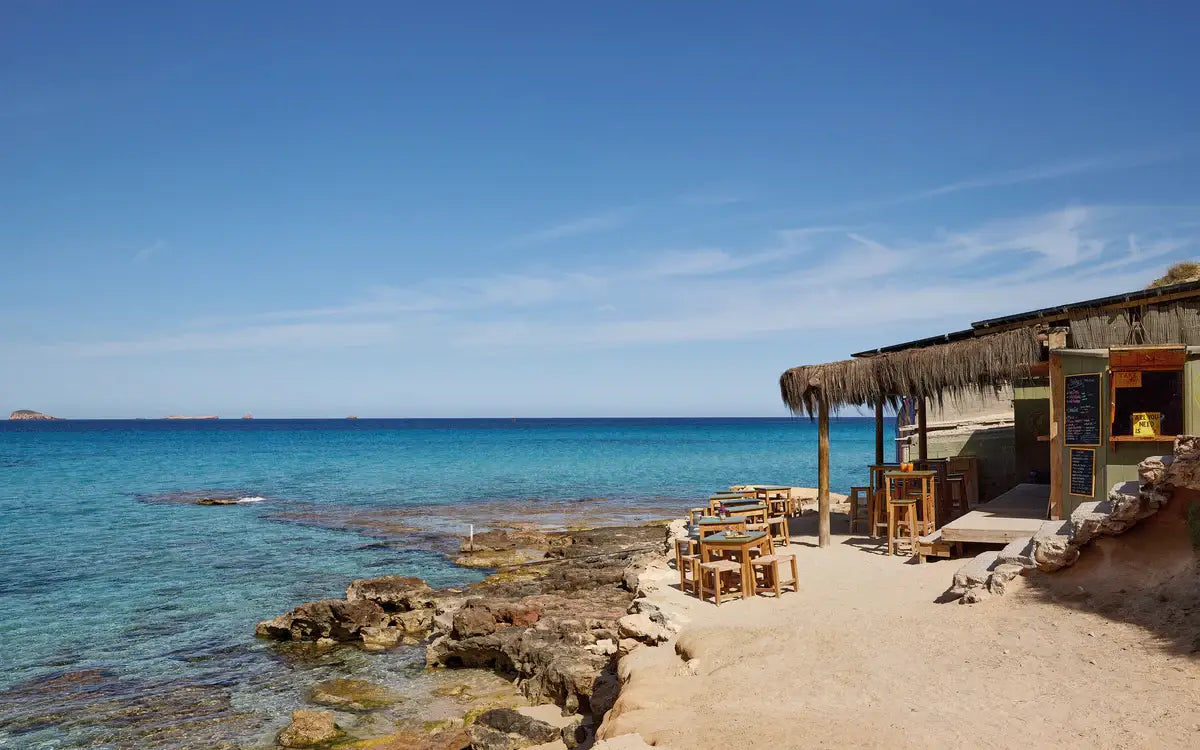 Outdoor wooden tables and chairs on a sandy beach by a thatched-roof bar evoke a tranquil coastal vibe, just like the scenes found in "The Coolest Ibiza" by Loft Publications.